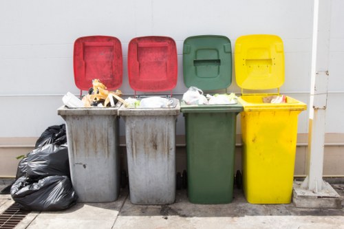 A row of four wheelie bins used for rubbish collection, positioned on a paved surface against a light grey exterior wall. From left to right, the bins are coloured grey, red, green, and yellow, each with their respective lids open or closed, revealing various waste materials inside such as plastic bags, cardboard, and miscellaneous refuse. To the left of the grey bins, there are multiple black plastic rubbish bags piled directly on the ground. The environment appears to be an outdoor driveway or yard area, typical for commercial waste collection in Neasden, with some dirt or debris visible on the paving and a drainage grate nearby. The overall scene reflects the typical setup for waste disposal and removal services, and it aligns with the services provided by Commercial Waste Removal Neasden in managing diverse rubbish types in the local area.