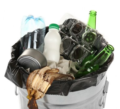 A metal waste bin lined with a black plastic bag is filled with various discarded items, including a crushed aluminum can, a worn-out brown leather shoe, a used white plastic bottle with a green cap, and empty green glass bottles. The bin is positioned outdoors on a paved surface, with a background of concrete or stone ground. The lighting appears natural, suggesting daytime, and the scene reflects typical rubbish collection imagery related to waste removal services such as those offered by Commercial Waste Removal Neasden, serving local businesses and residents in the Neasden area within postcode NW10. The varied materials within the bin—including metal, glass, plastic, and organic debris—highlight waste types commonly dealt with during rubbish removal processes in London suburbs. The arrangement and visible textures emphasize the typical contents encountered when clearing commercial or household waste, supporting the description's relevance to waste management activities in the local area.