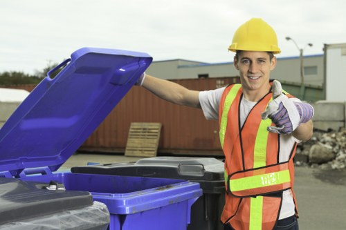 A young male waste management worker wearing a yellow safety helmet, a high-visibility orange and yellow vest, and purple gloves stands outdoors in an industrial or commercial area. He is smiling and giving a thumbs-up gesture while holding the lid of a blue wheeled rubbish bin that is open, revealing black waste containers inside. The background features a paved surface with stacks of construction and waste materials, a wooden pallet, and some industrial buildings under a cloudy sky. The scene reflects professional rubbish collection and waste removal services, consistent with the offerings of Commercial Waste Removal Neasden, operating within the local commercial waste management sector.