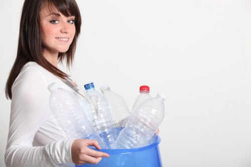 A young woman with shoulder-length brown hair and a friendly expression is holding a blue plastic bin filled with used transparent plastic bottles, some with coloured caps, set against a plain white background. The bottles vary in size and are visibly crumpled and partially filled with liquid residue, indicating they are ready for recycling or disposal. She grips the sides of the bin firmly with both hands, supporting it at waist level. The scene is well-lit, emphasizing the clarity of the bottles and the smooth surface of the bin. This image contextually relates to rubbish collection or waste management services, such as those offered by Commercial Waste Removal Neasden, focusing on recycling waste materials like plastic bottles. The setting appears to be a simple indoor environment, free from other objects or clutter, with clear attention on the bottles and the woman's interaction with them, illustrating responsible waste handling in a straightforward, professional manner.