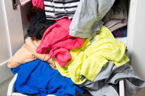 A close-up of an open metal wardrobe drawer filled with piled laundry, including various clothing items such as a bright pink shirt, a yellow garment, a blue piece of clothing, and several grey, black, and beige pieces, all made of different fabrics with visible textures. The drawer is partially pulled out, revealing the mixed clothes stacked and spilling over the edges. The background shows part of a white interior with additional closet shelving or storage units. This scene depicts cluttered laundry, which could be part of domestic waste or rubbish collection in a residential setting, relevant to rubbish removal services for Neasden or nearby London areas. The overall environment is indoors, with natural lighting highlighting the multicoloured clothing textures.